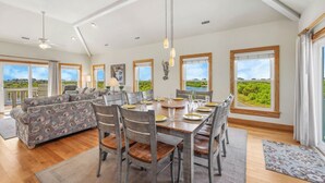 A spacious living room with a dining area, large windows offering scenic views, and a ceiling fan. - Soundside Dog Friendly Home w/ Private Pool, Hot Tub, Elevator, and Game Room (Avon)