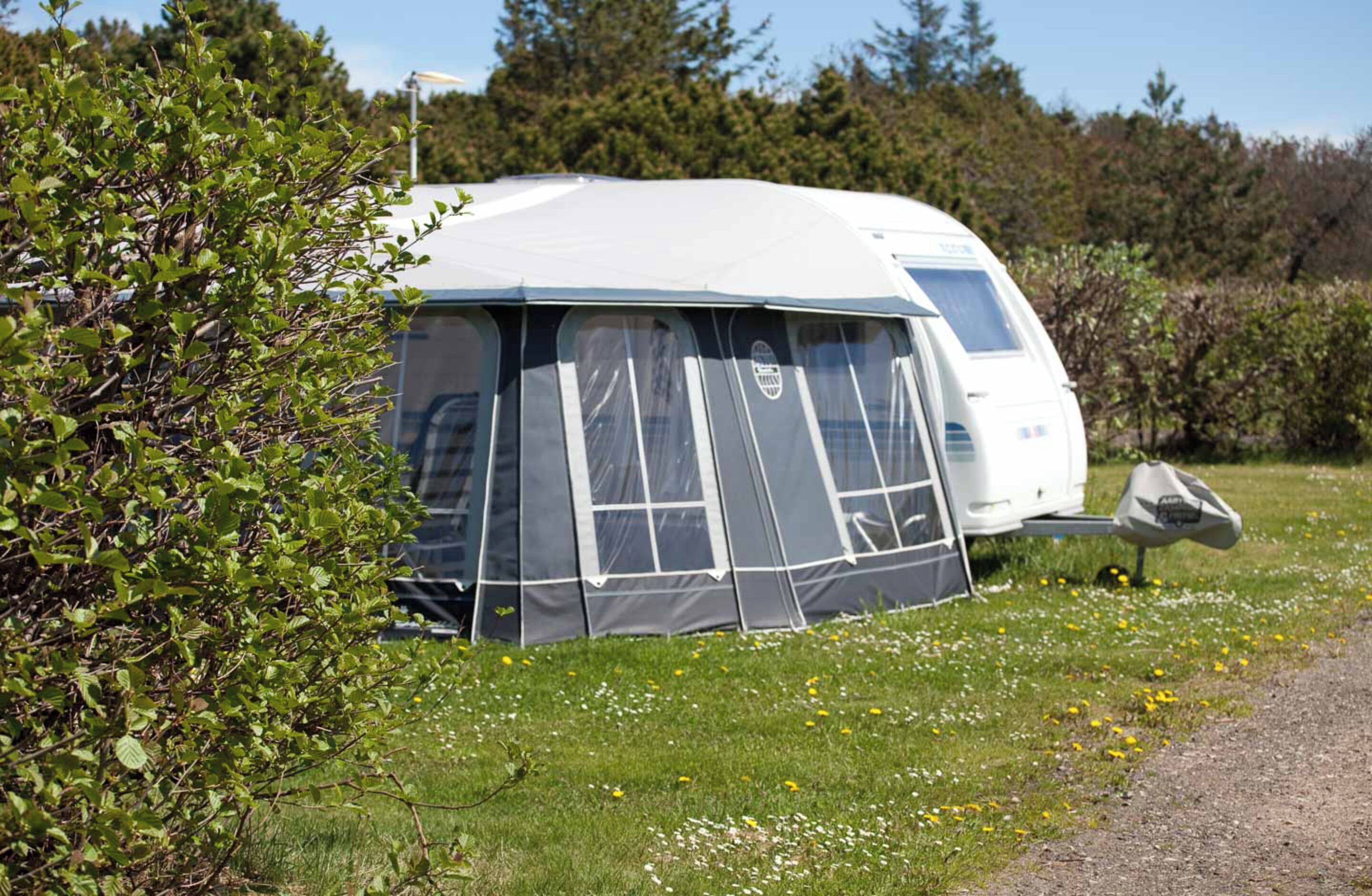 Un camping-car blanc, dont les fenêtres avant et latérales sont recouvertes, est garé sur une pelouse, avec un buisson au premier plan.