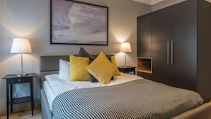 Et moderne soveværelse med en stor seng, to sengeborde med lamper, en mørk garderobe og et indrammet abstrakt maleri på væggen.