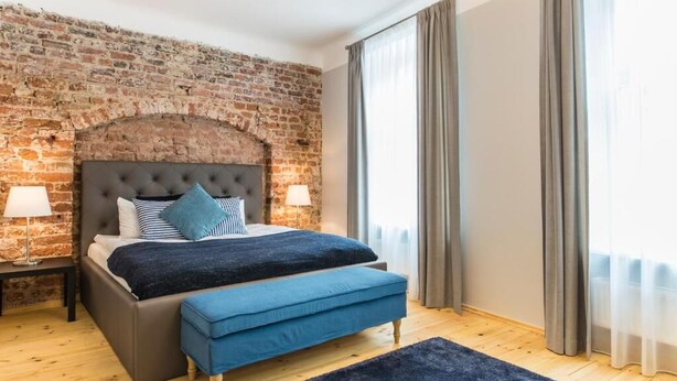 Ein Schlafzimmer mit Backsteinwand, einem gesteppten Kopfteil, einem Bett mit blauer Bettwäsche, einer blauen Bank und einem Nachttisch mit Lampe.