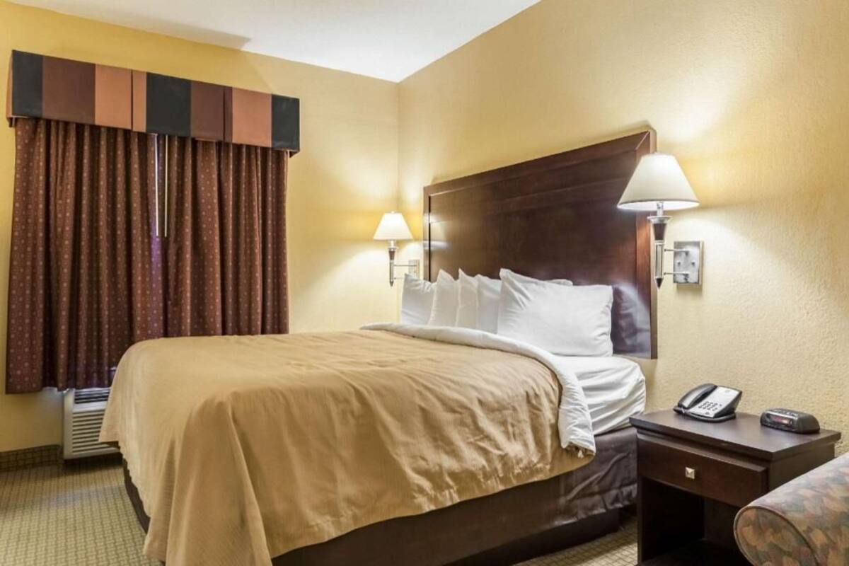 A hotel room with a bed, two bedside lamps, a nightstand with a phone, and a window with curtains.