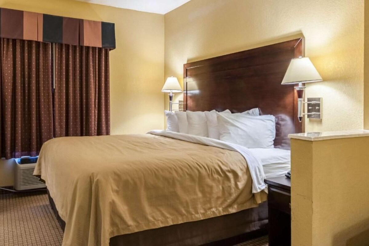Ein Hotelzimmer mit einem großen Bett, zwei Nachttischlampen, einem hölzernen Kopfteil und einem Fenster mit Vorhängen.