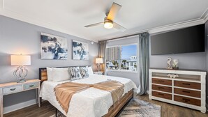 Une chambre à coucher avec un lit, des tables de chevet, un ventilateur de plafond, une télévision et une vue sur l’extérieur.