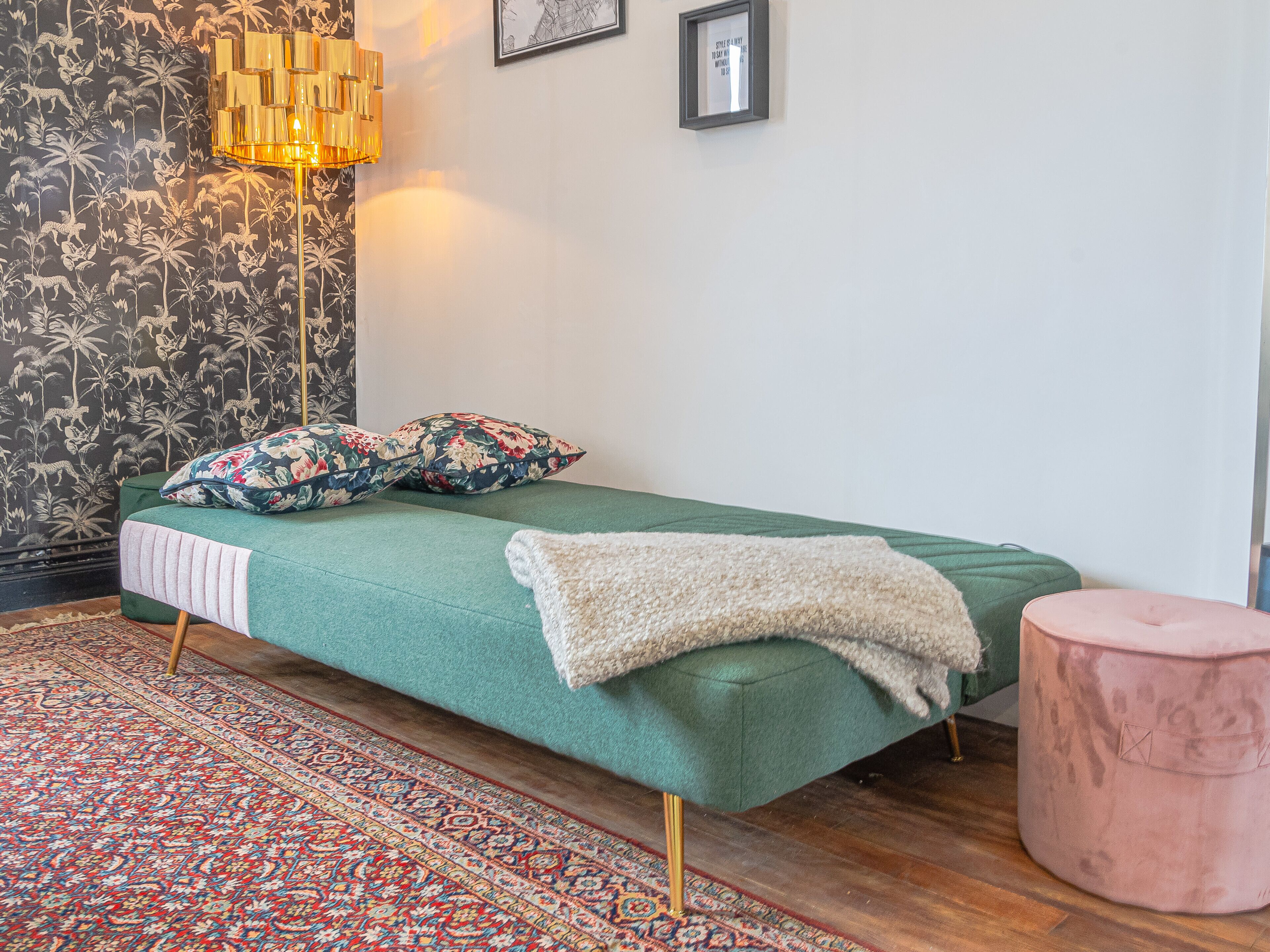 A room with a green bed, floral patterned wallpaper, a floor lamp, and a patterned rug.