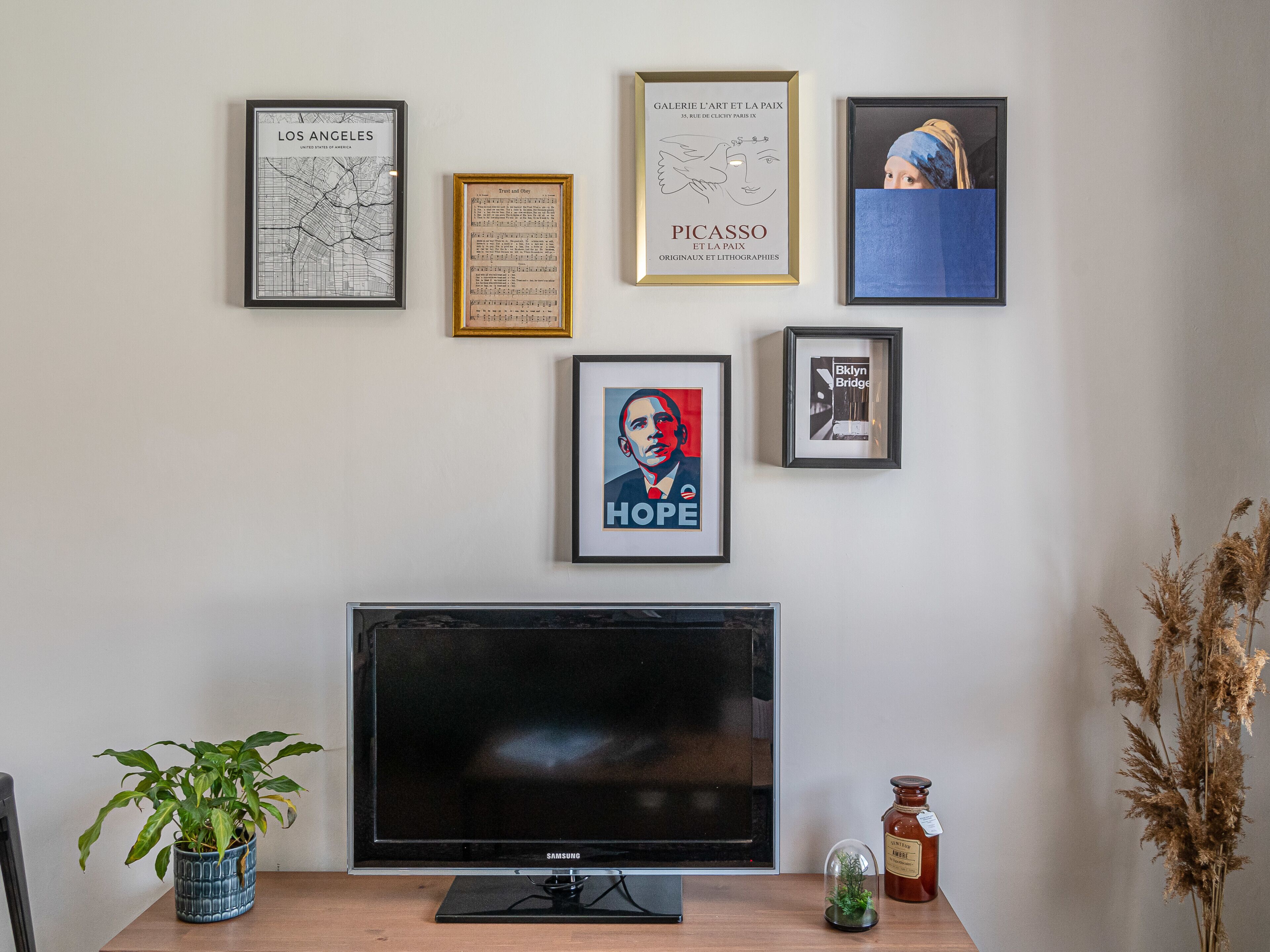 Een muur met ingelijste kunstwerken, een televisie, een potplant en een potje jam.