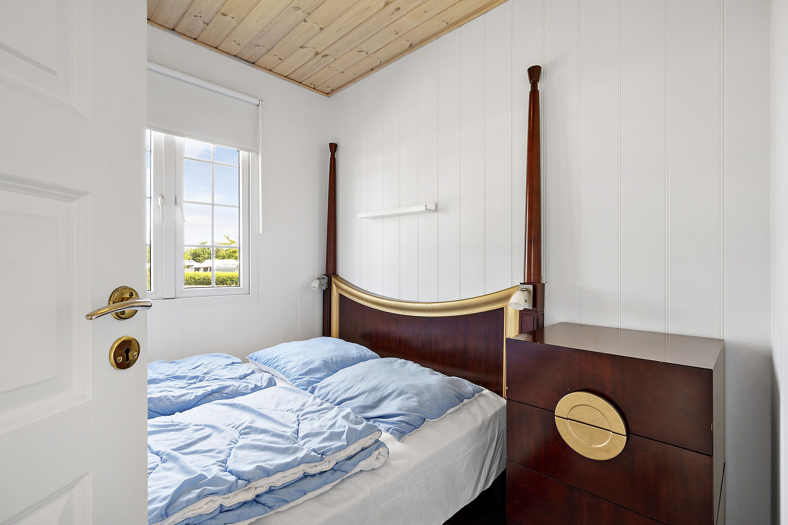 Une chambre à coucher avec une tête de lit en bois, un lit avec une literie bleue, une table de chevet et une fenêtre donnant sur un paysage verdoyant.