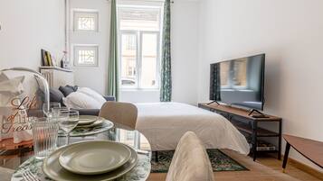 A hotel room with a bed, a TV, a dining table with plates and glasses, a chair, and a window with curtains.