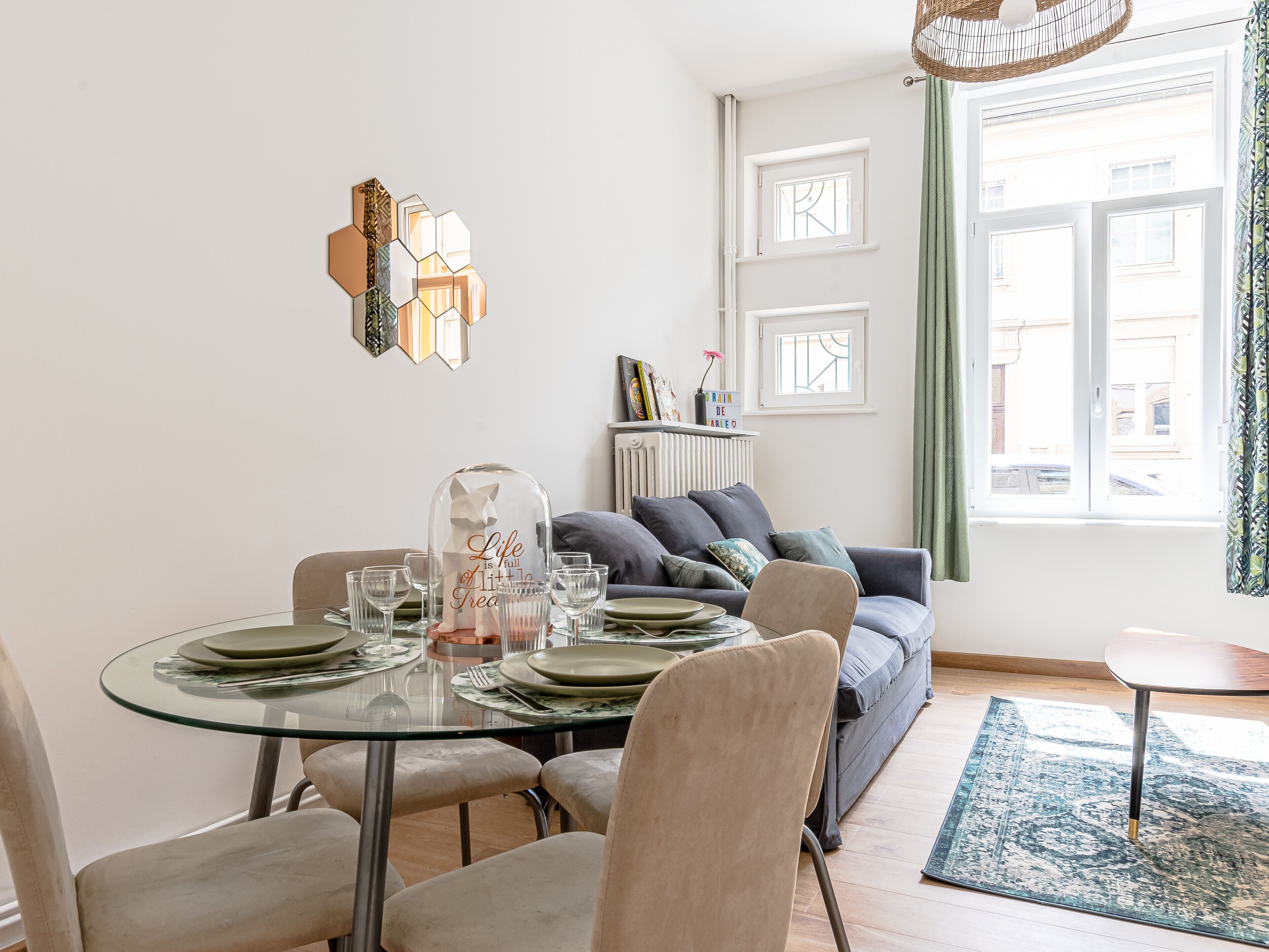 Un coin repas avec une table en verre dressée pour quatre personnes, un canapé avec des coussins et une vue sur la rue par une fenêtre.