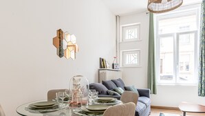 A dining area with a glass table set for four, a sofa with cushions, and a view of the street through a window. - Rock in Share - Le Grain de Sable (Thionville)