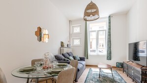 A modern living room with a glass dining table, grey sofa, and a television on a wooden cabinet. - Rock in Share - Le Grain de Sable (Thionville)