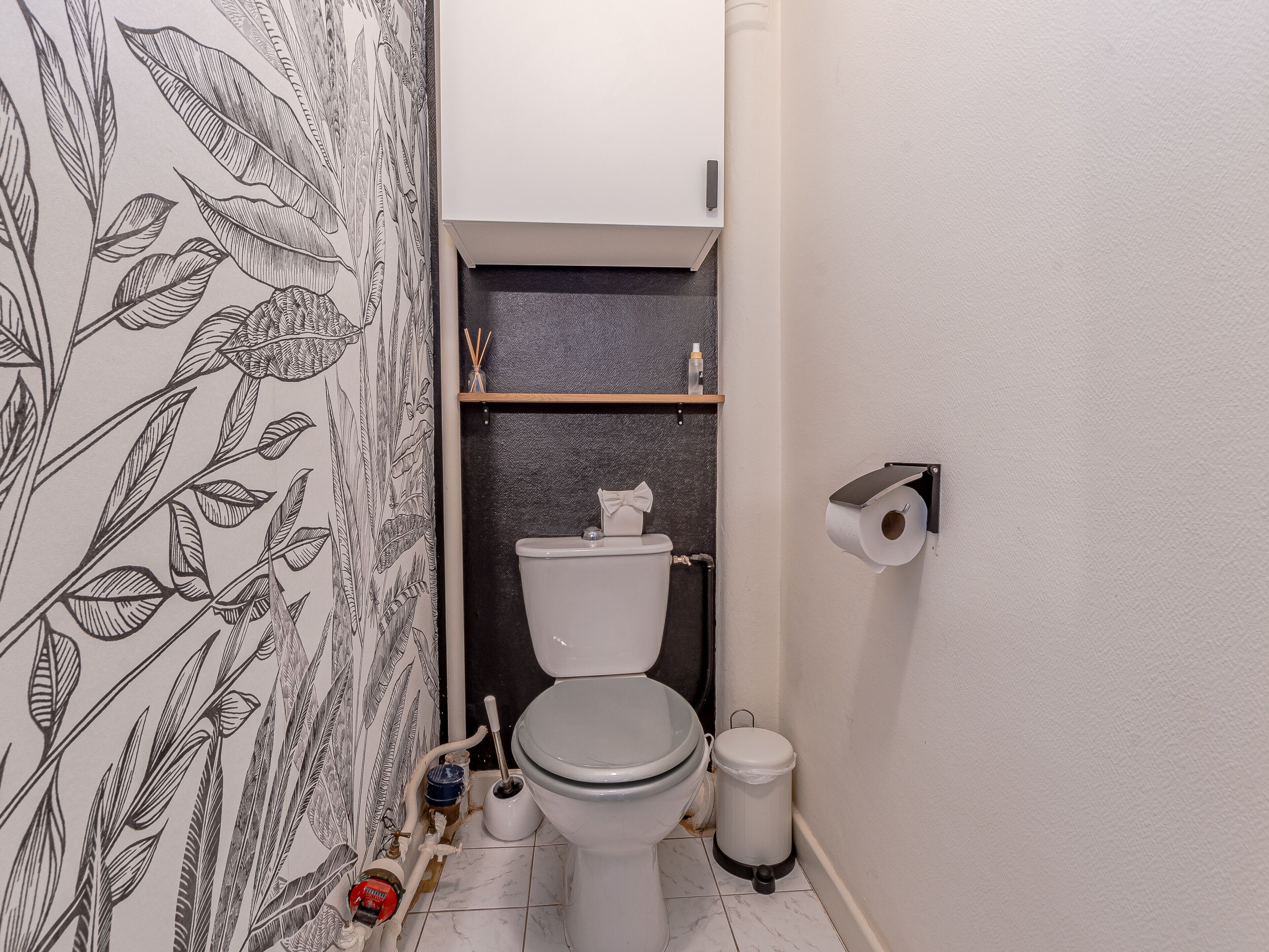 A bathroom with a toilet, a trash can, a toilet paper holder, and a shelf with a bottle and reed diffuser.