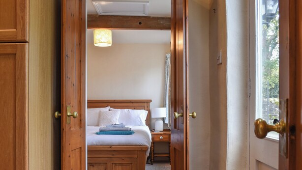 A wooden door leading into a bedroom with a bed, bedside table, lamp, and a view of the outdoors through a window.