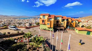 Un edificio frente al mar con varios balcones, zona de estacionamiento y vistas a la montaña.