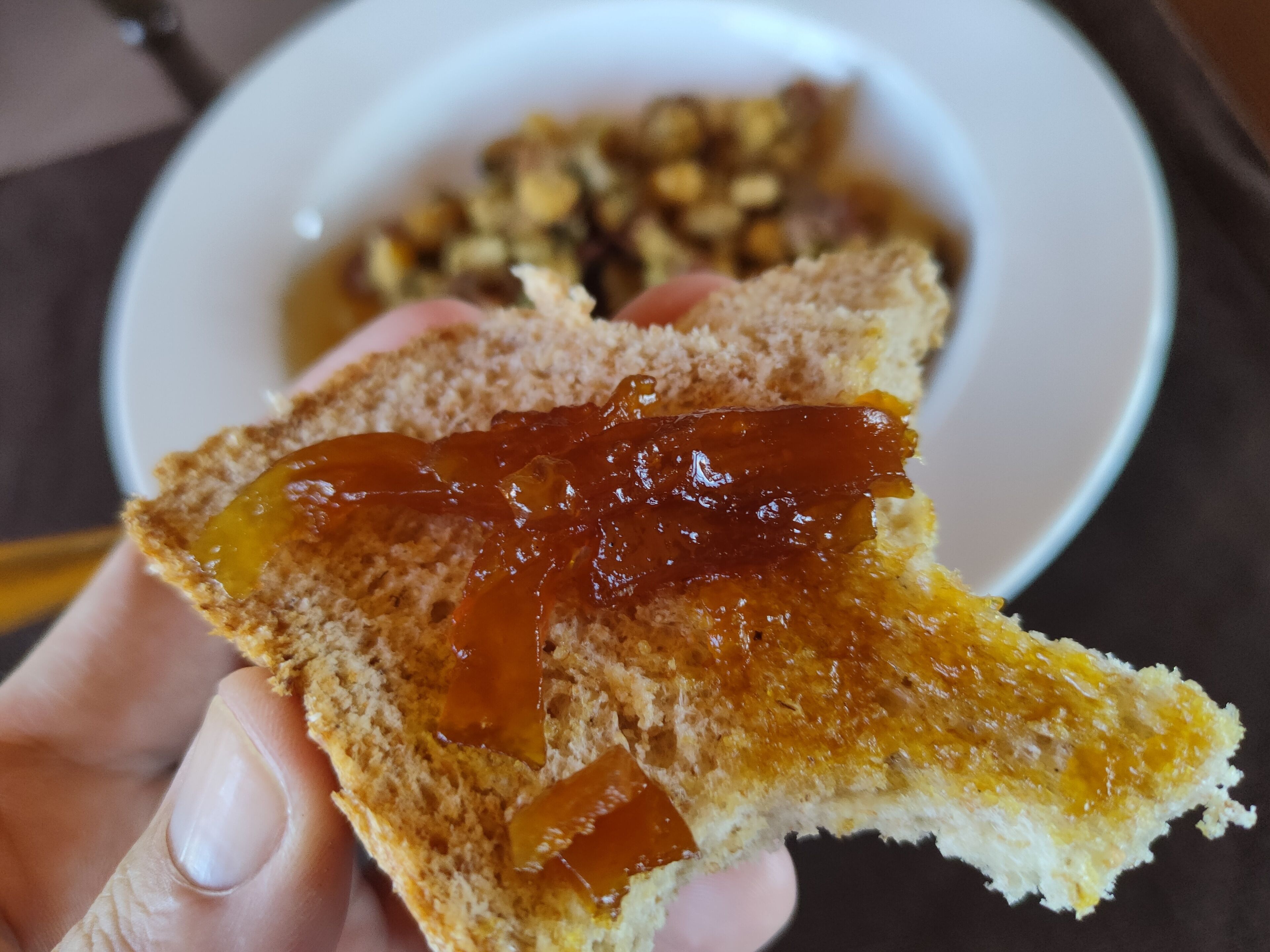 Une tranche de pain grillé tartinée de confiture, tenue dans une main, avec un bol de noix en arrière-plan.