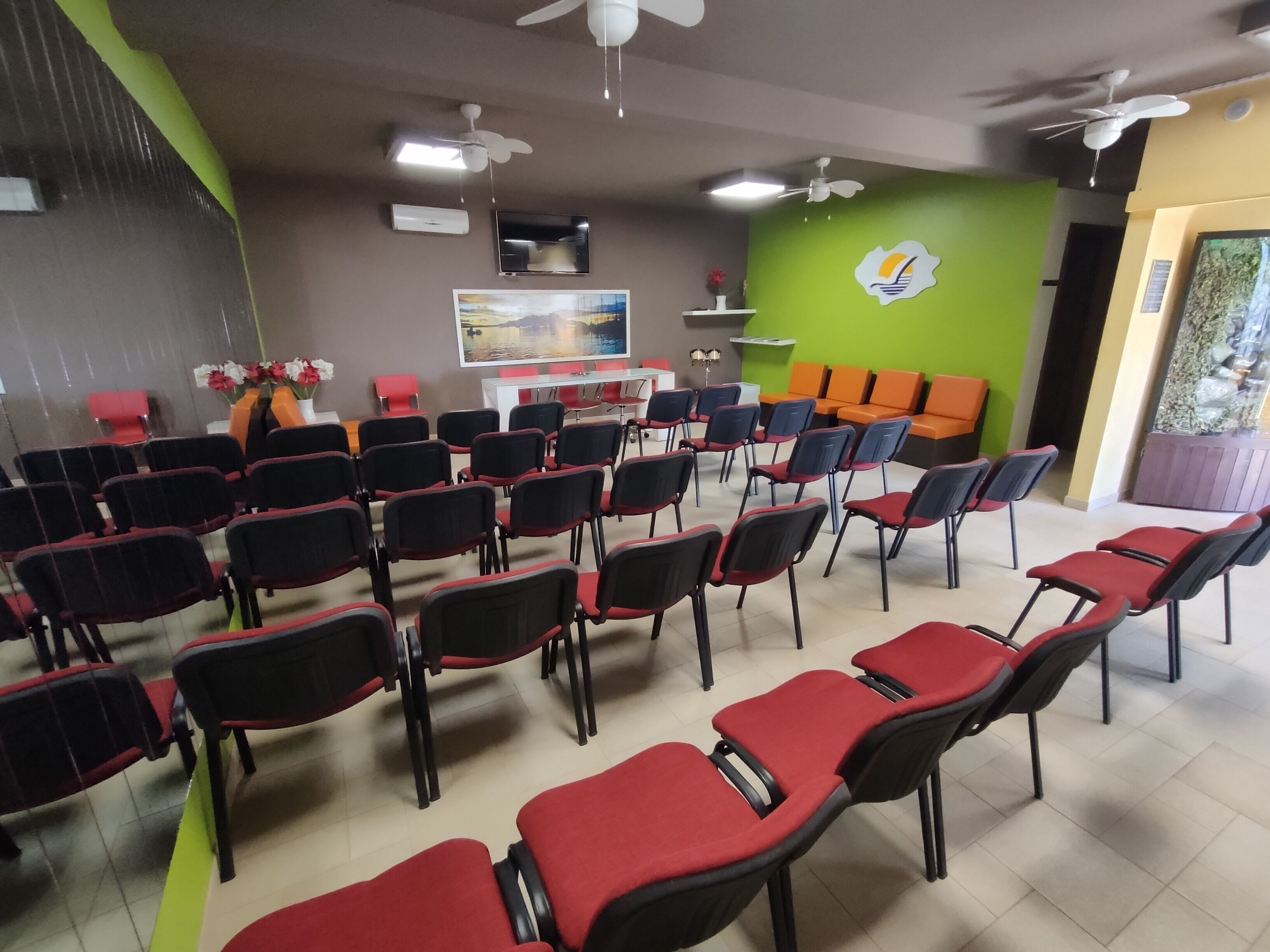Un salón de conferencias con filas de sillas, un televisor, ventiladores de techo y una pared verde con un logotipo.