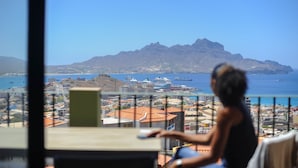 Una persona sentada a una mesa con vista a una ciudad costera, incluyendo montañas y un cuerpo de agua.
