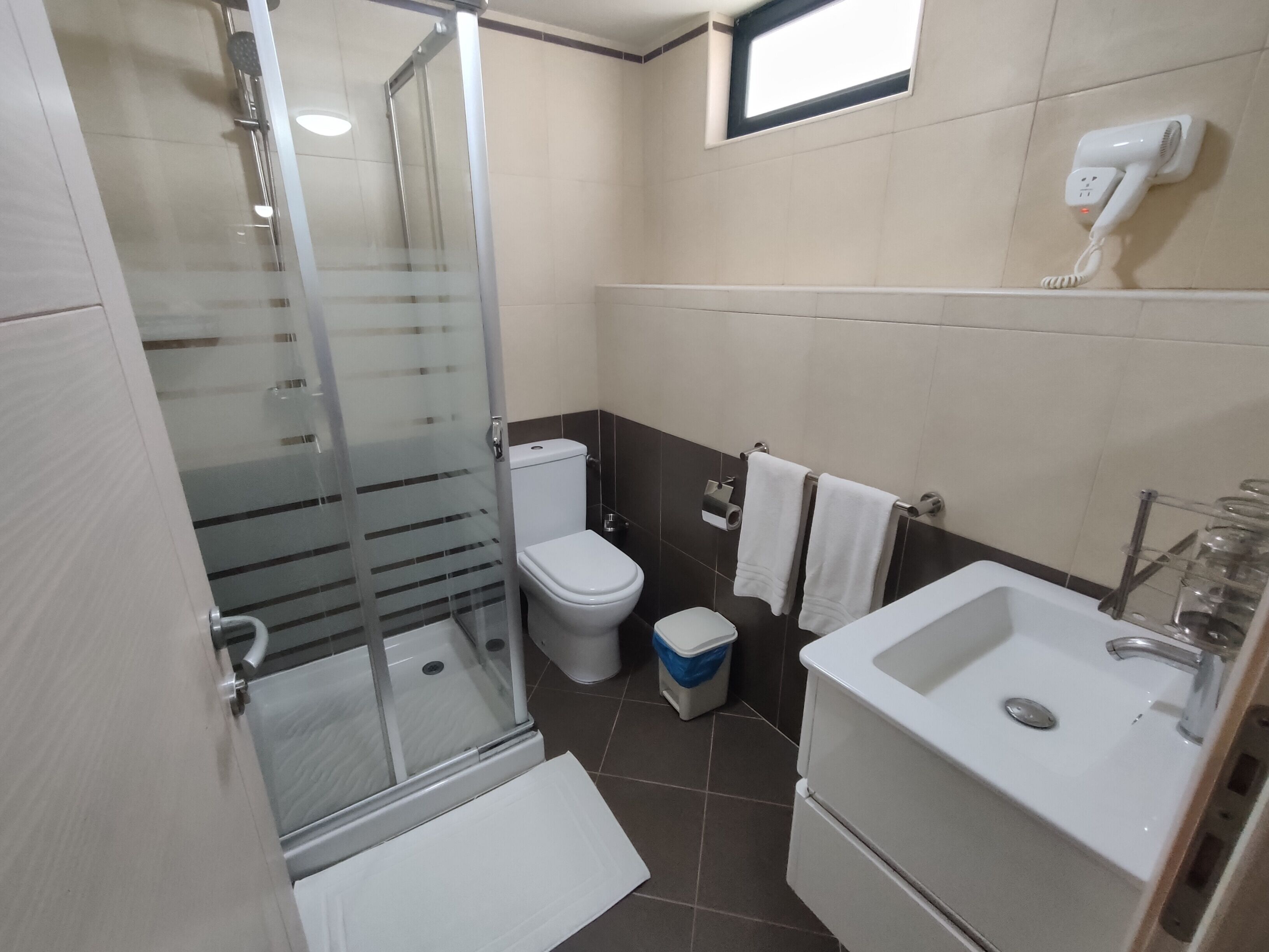 Egy modern fürőszoba, melyben üvegfalas zuhanyzó, WC, mosdókagyló és a falra szerelt hajszárító található.