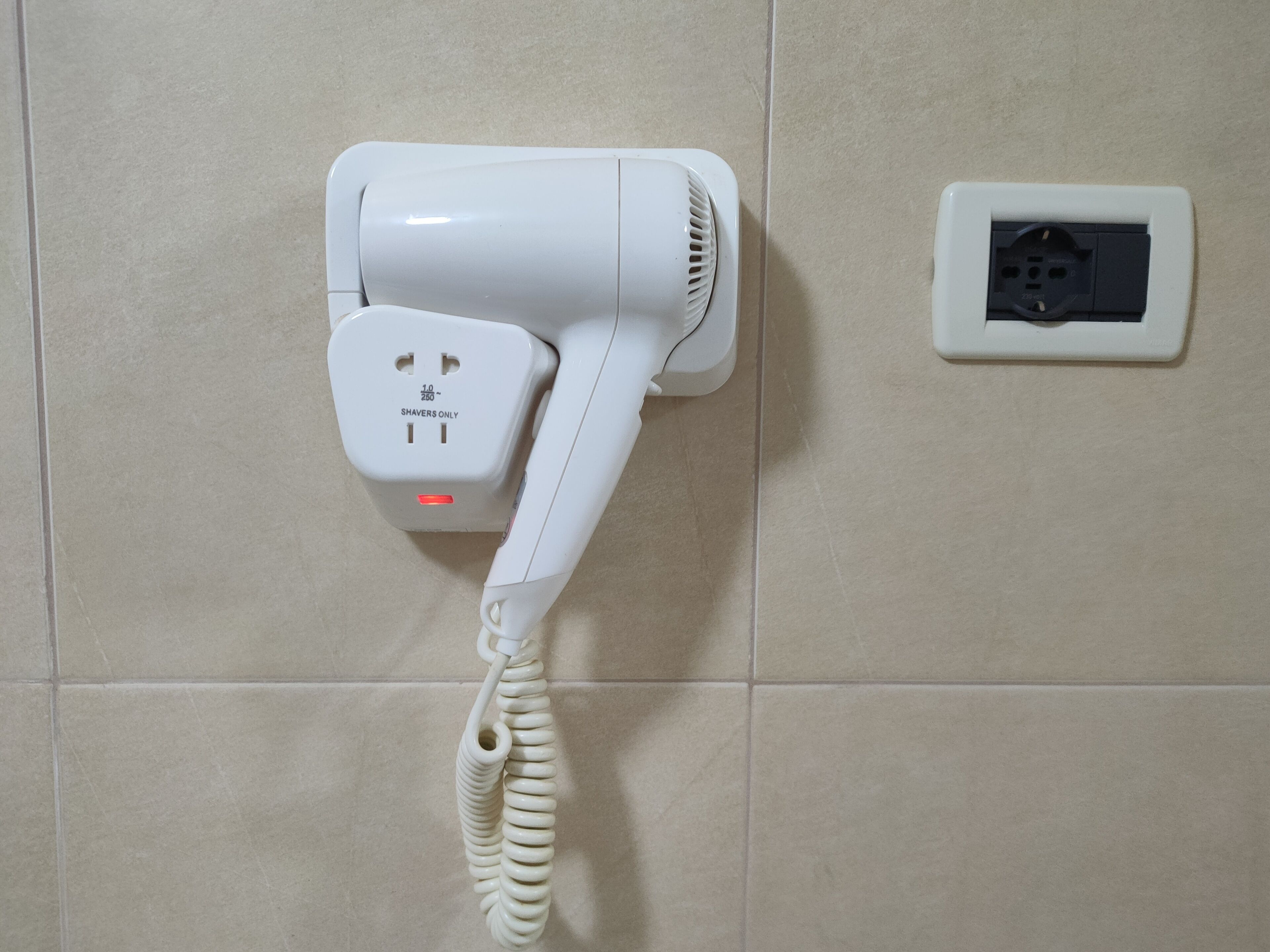 Um secador de cabelo fixado numa parede revestida de azulejos, ligado a uma tomada.