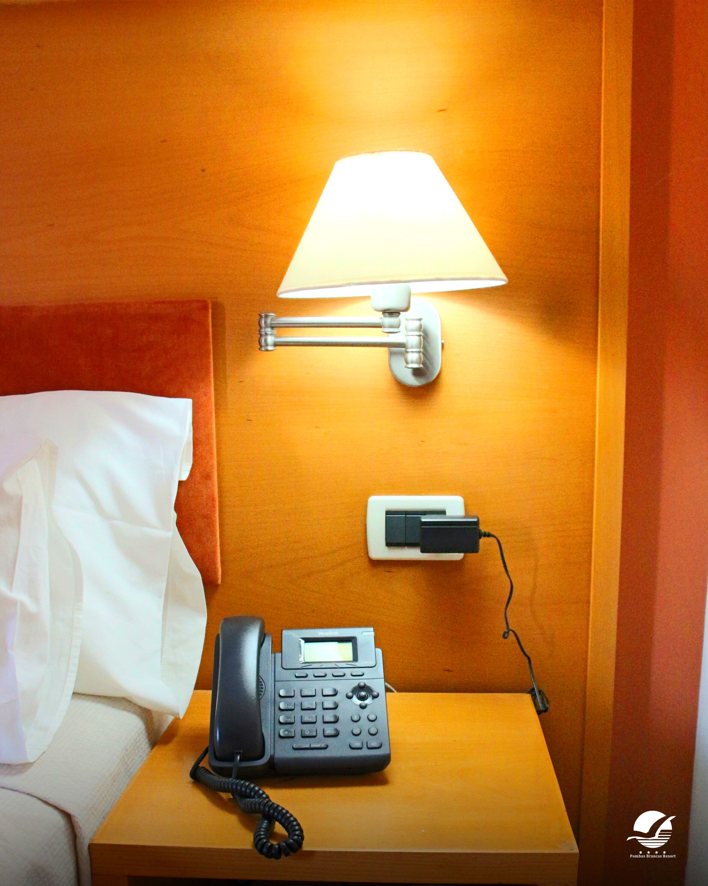 Habitación de hotel con una mesita de noche de madera, una cama blanca con un cojín blanco, un teléfono negro, una lámpara de pared y un enchufe.