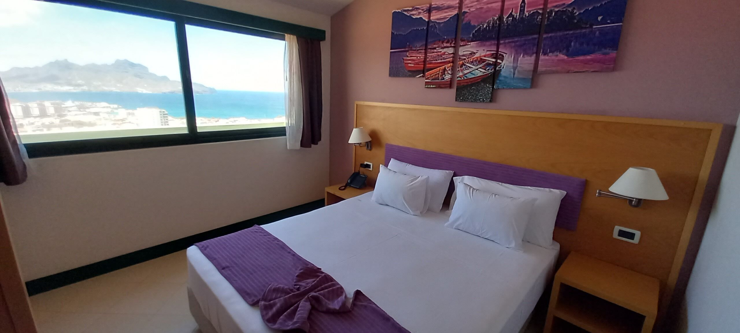 A hotel room with a large bed, two bedside tables, a nightstand, a window with a view of the sea, and a painting on the wall.