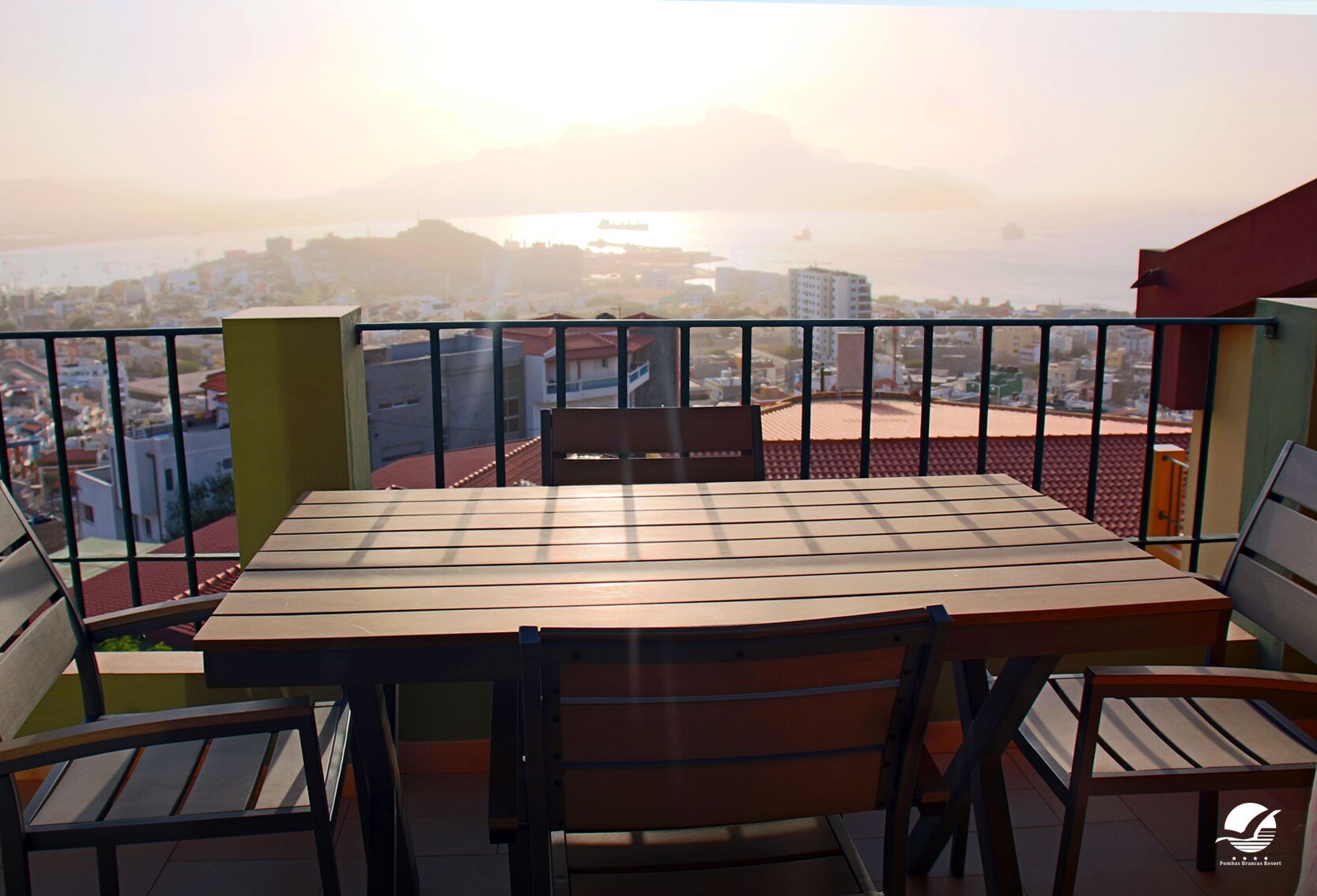 A balcony with a table and chairs, offering a view of a cityscape and mountains in the distance.