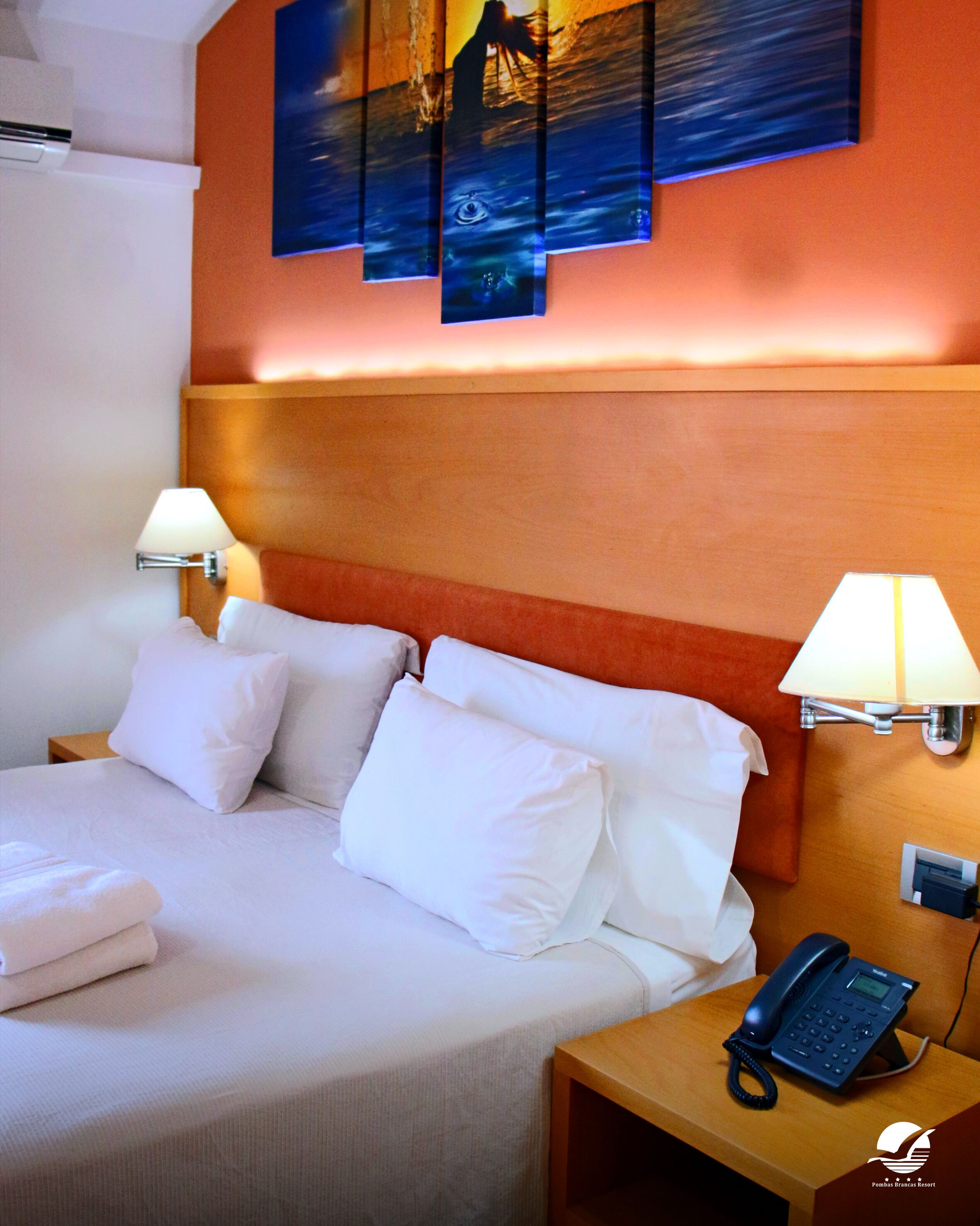 Una habitación de hotel con una cama, dos lámparas de noche, un teléfono en una mesita y un cuadro en la pared.