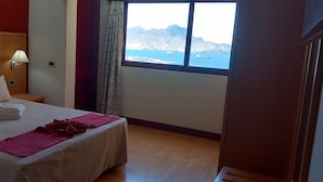 Habitación de hotel con cama, mesita de noche, lámpara, ventana con vistas al mar y a las montañas, y una puerta.