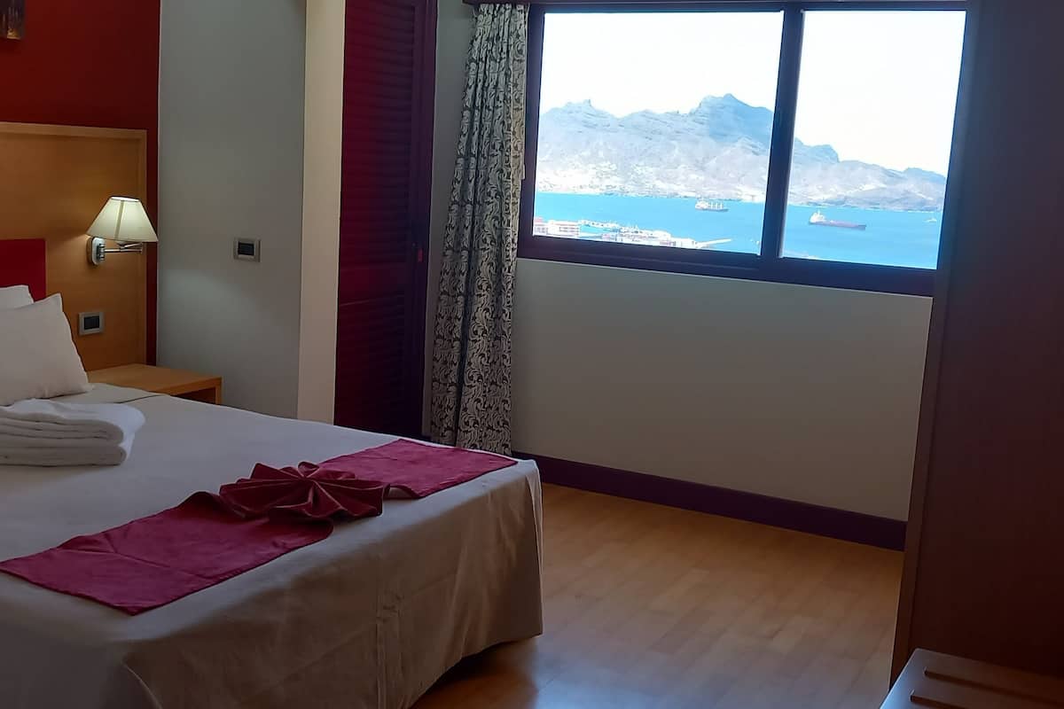 A hotel room with a bed, a nightstand, a lamp, a window with a view of the sea and mountains, and a door.