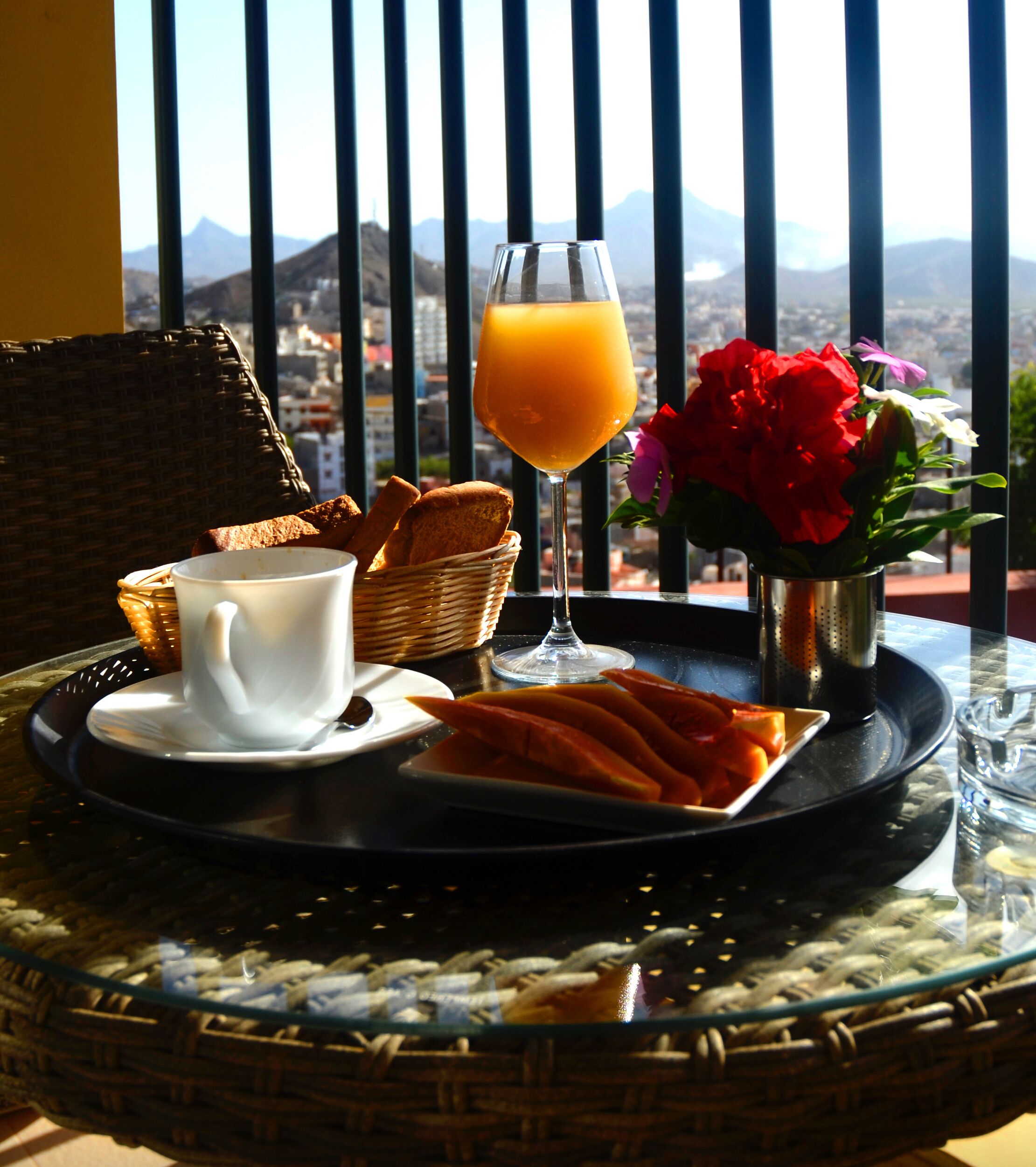 Ein Frühstücksbrett mit Brot, einer Tasse Kaffee, einem Glas Orangensaft und einer Fruchtplatte, auf einem Tisch mit Bergblick.