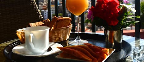 Bandeja de desayuno con pan, una taza de café, un vaso de zumo de naranja y un plato de fruta, sobre una mesa con vistas a las montañas.