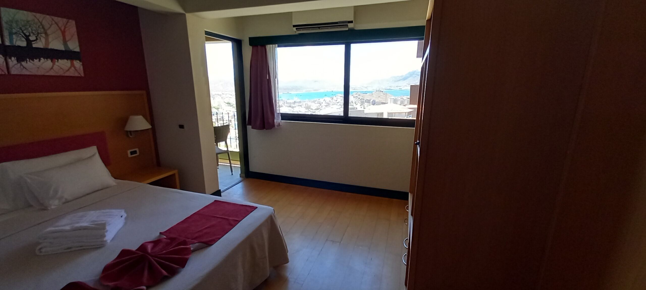 Une chambre d’hôtel avec un lit, une table de chevet, une fenêtre donnant sur un paysage urbain et un balcon avec une chaise.