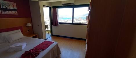 Une chambre d’hôtel avec un lit, une table de chevet, une fenêtre donnant sur un paysage urbain et un balcon avec une chaise.