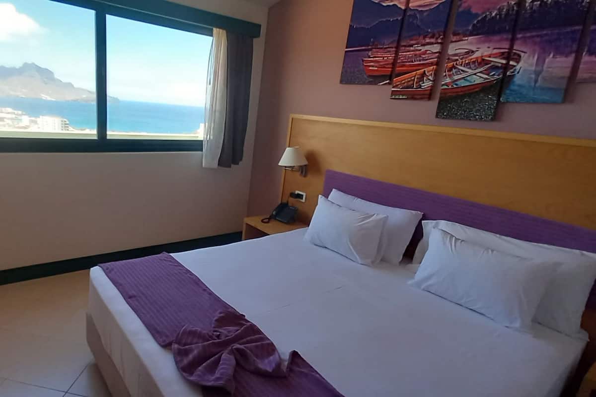 A hotel room with a bed, a nightstand, a lamp, and a painting of boats on the wall.