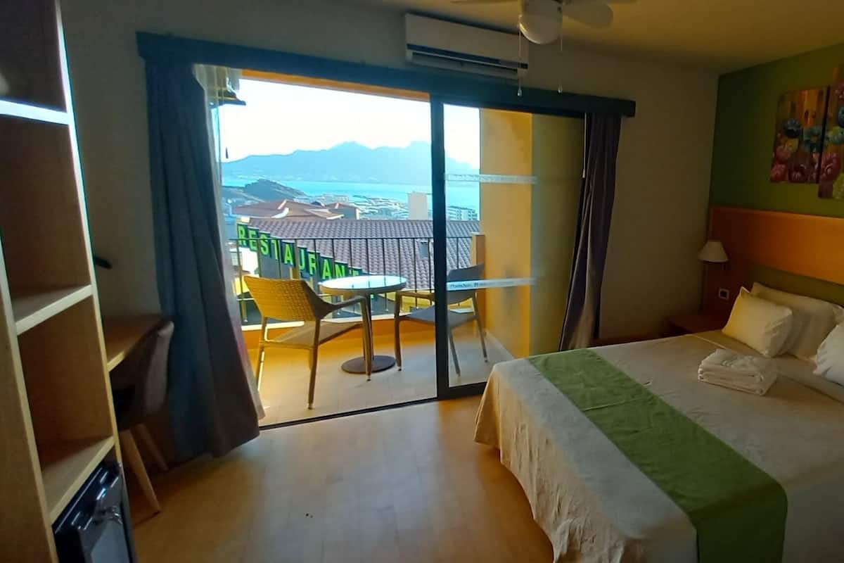 A hotel room with a bed, a balcony with a table and chairs, and a view of the sea.
