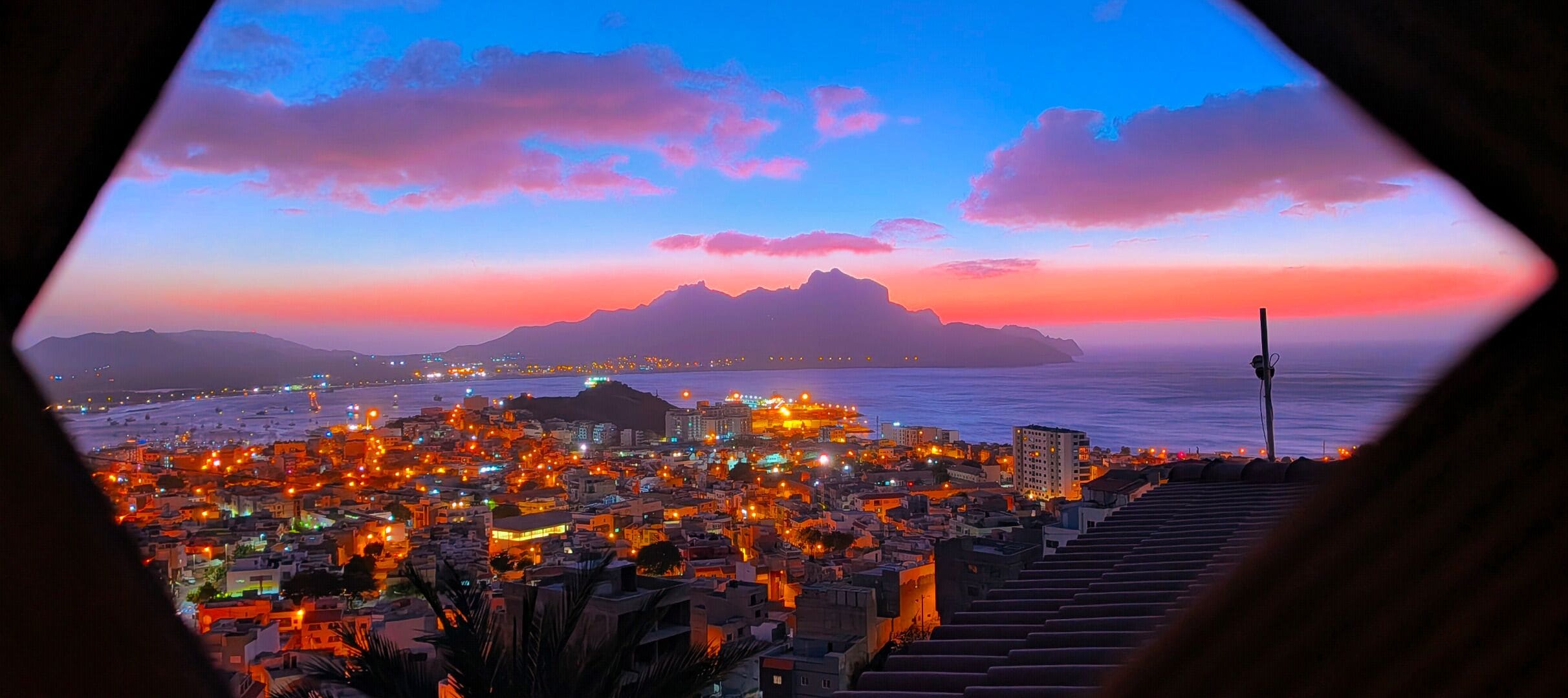 Une vue sur une ville au crépuscule, avec une montagne en arrière-plan et la mer en perspective.
