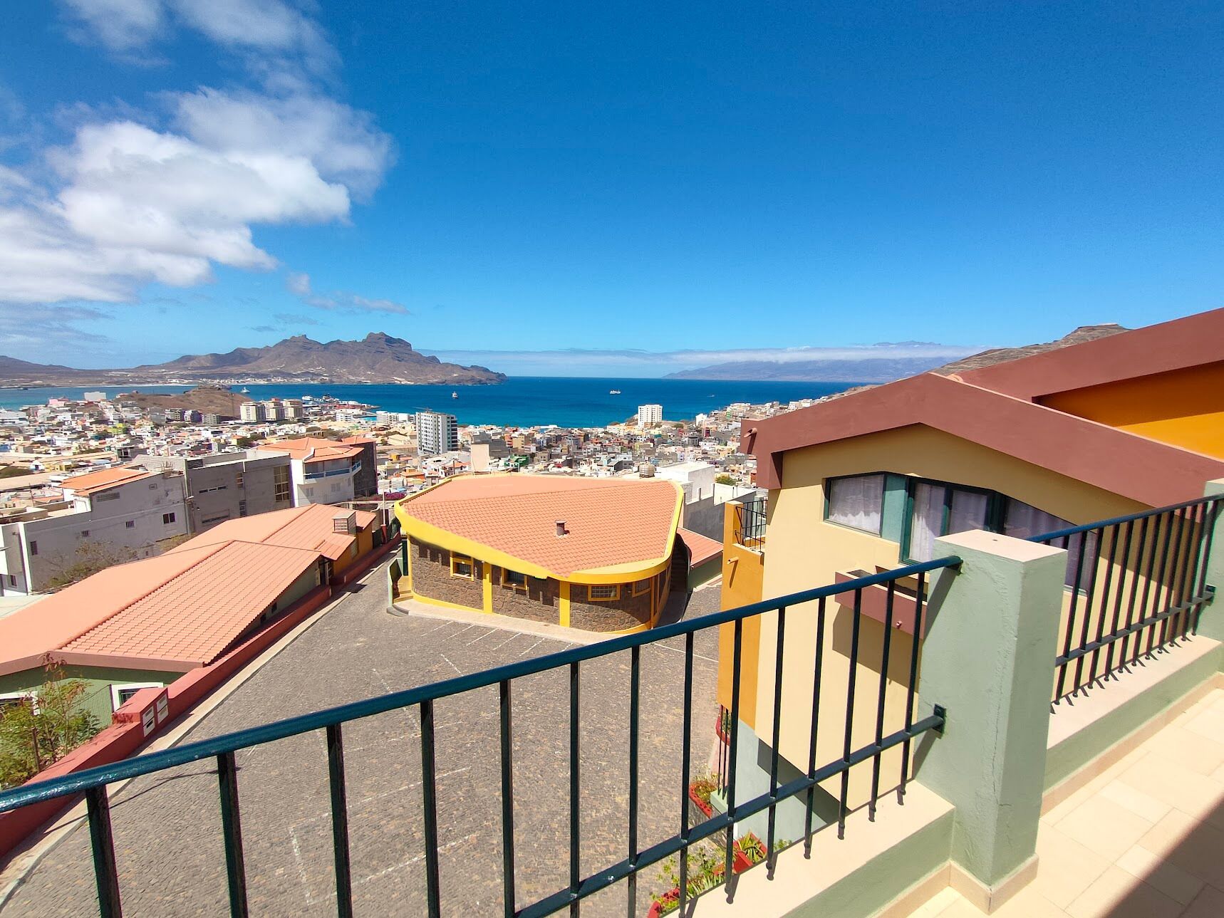 A high vantage point view of a coastal city with a clear blue sky, a prominent mountain in the distance, and a view of the sea.