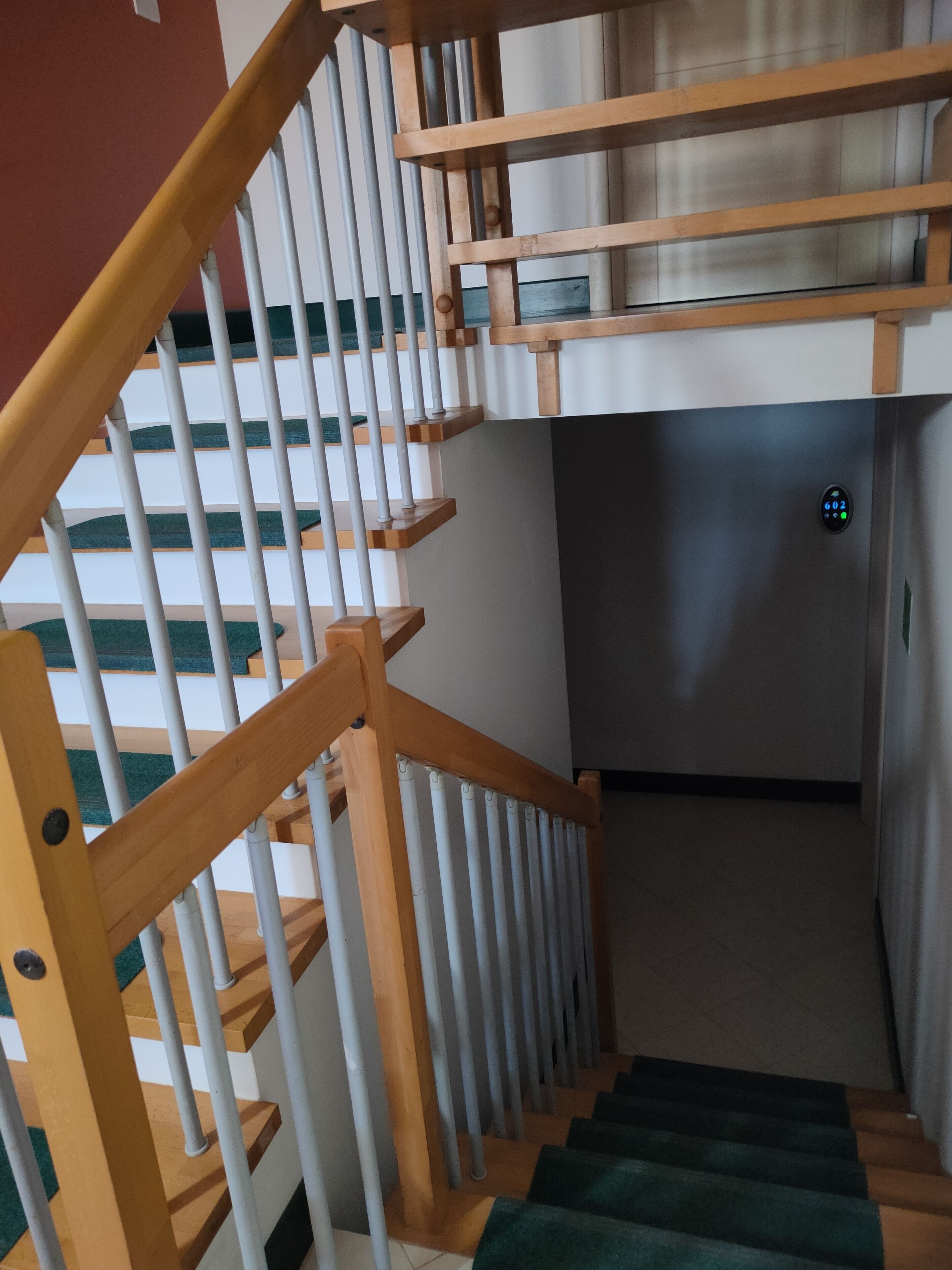 Un escalier avec des rampes en bois et des balustrades blanches, menant à un palier où se trouve un panneau de commande numérique pour l’ascenseur.