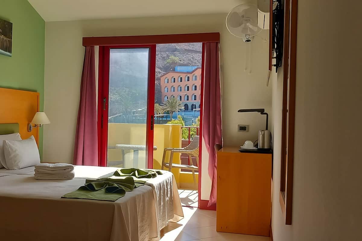 A hotel room with a bed, a table, a chair, a balcony with a view of buildings, and a window with curtains.