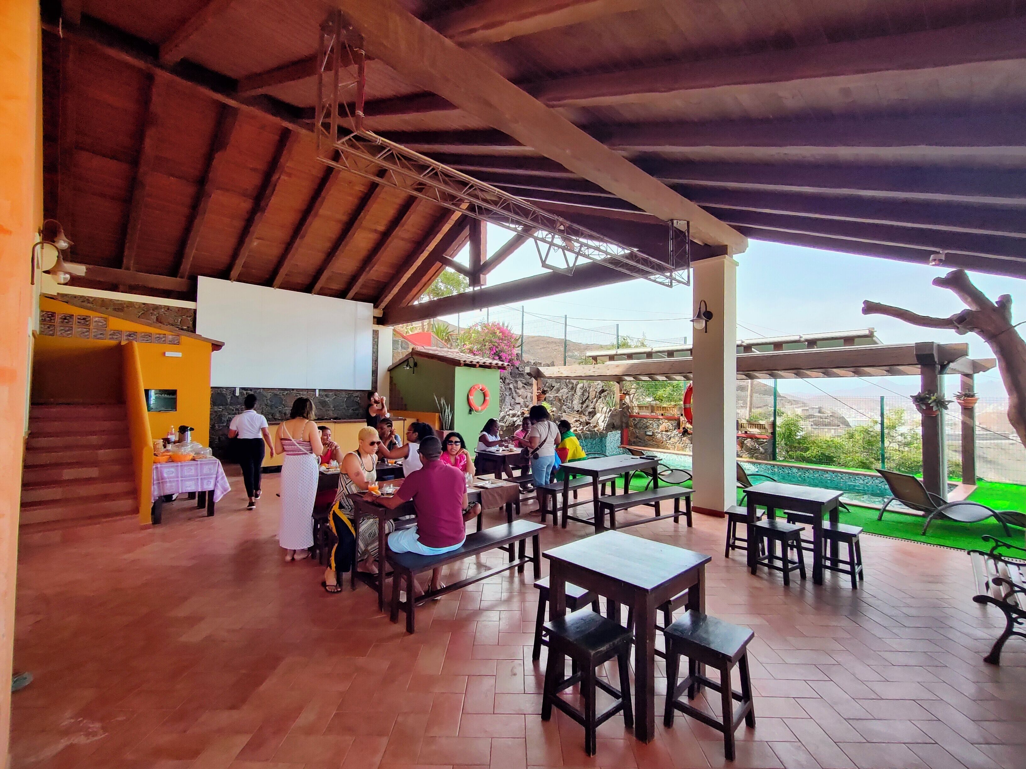 An outdoor dining area with wooden tables and benches, a covered roof, and a view of a pool and greenery.