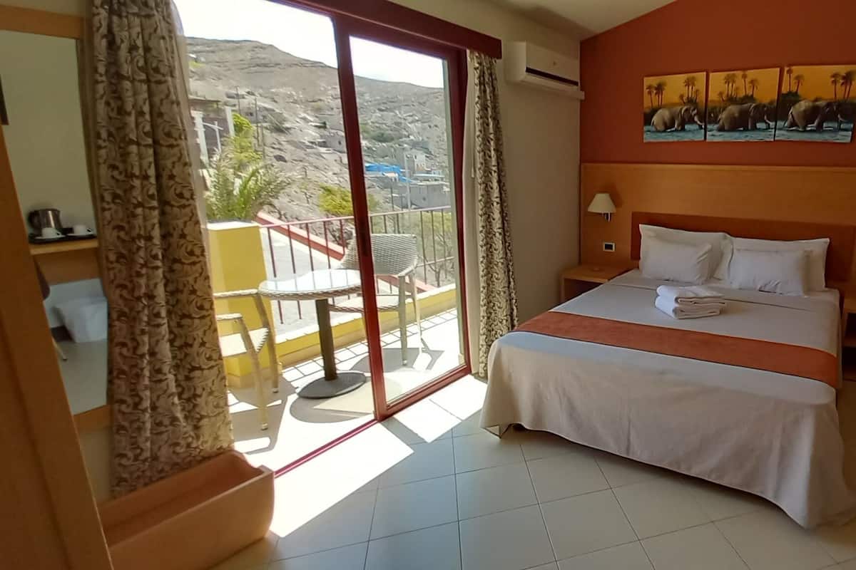 A hotel room with a bed, two bedside tables, a balcony with a chair, and a painting on the wall.