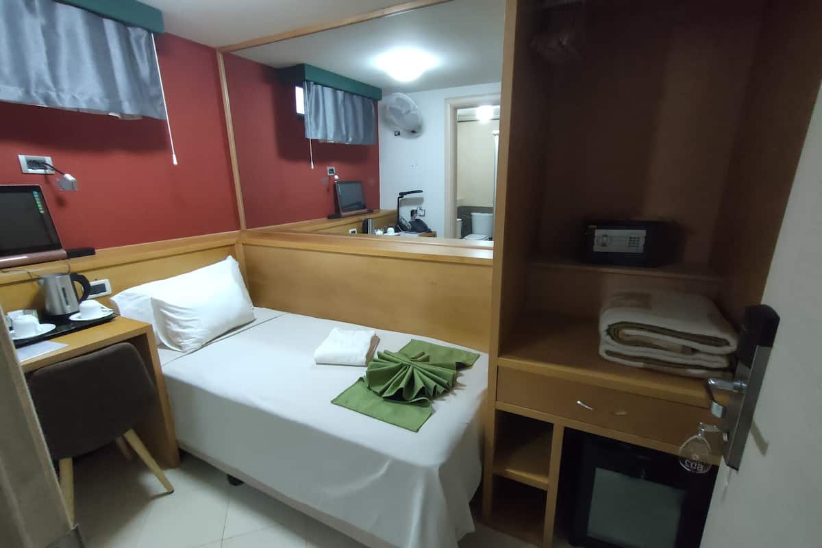 A hotel room with a bed, a desk with a computer, a mirror, a small shelf with towels, and a wall-mounted air conditioner.