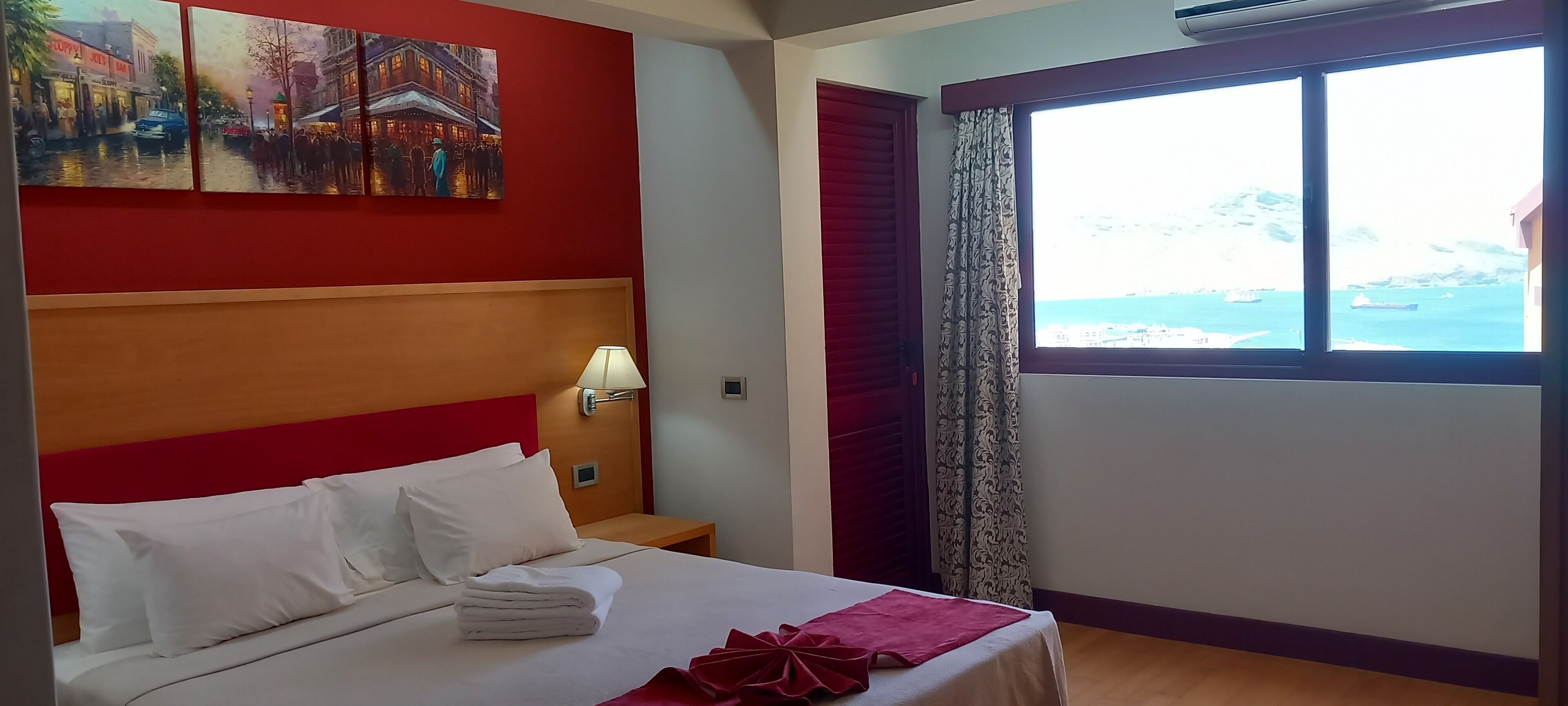 Una habitación de hotel con cama, mesita de noche, lámpara, ventana con vista al mar y un cuadro en la pared.