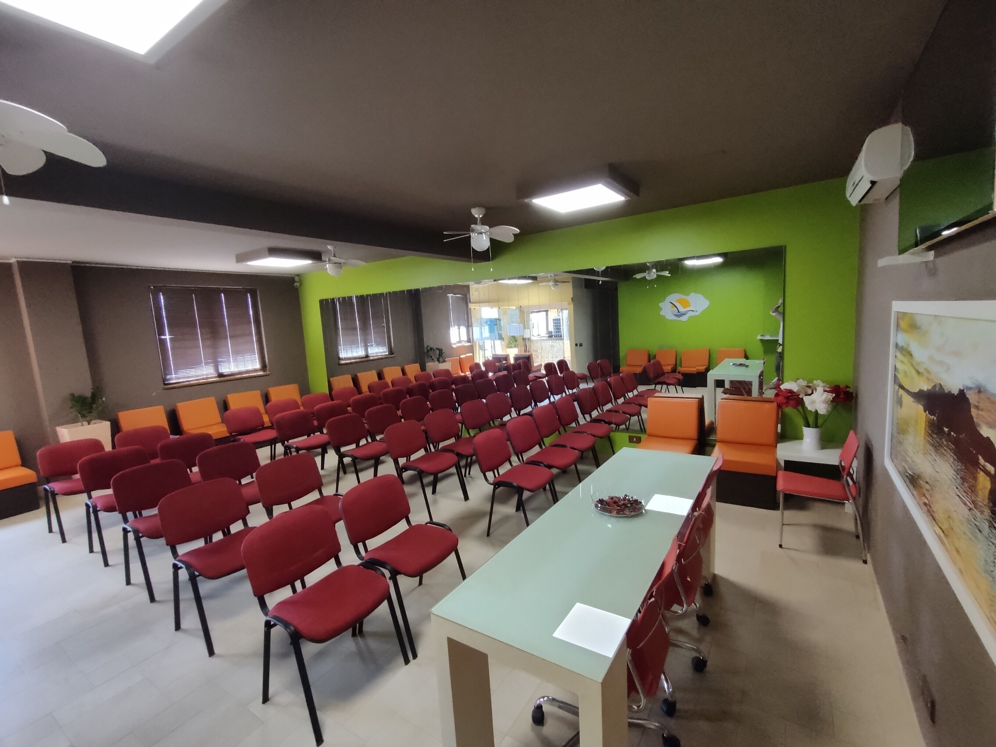 A conference room with rows of red chairs, a long table, green walls, and ceiling fans.