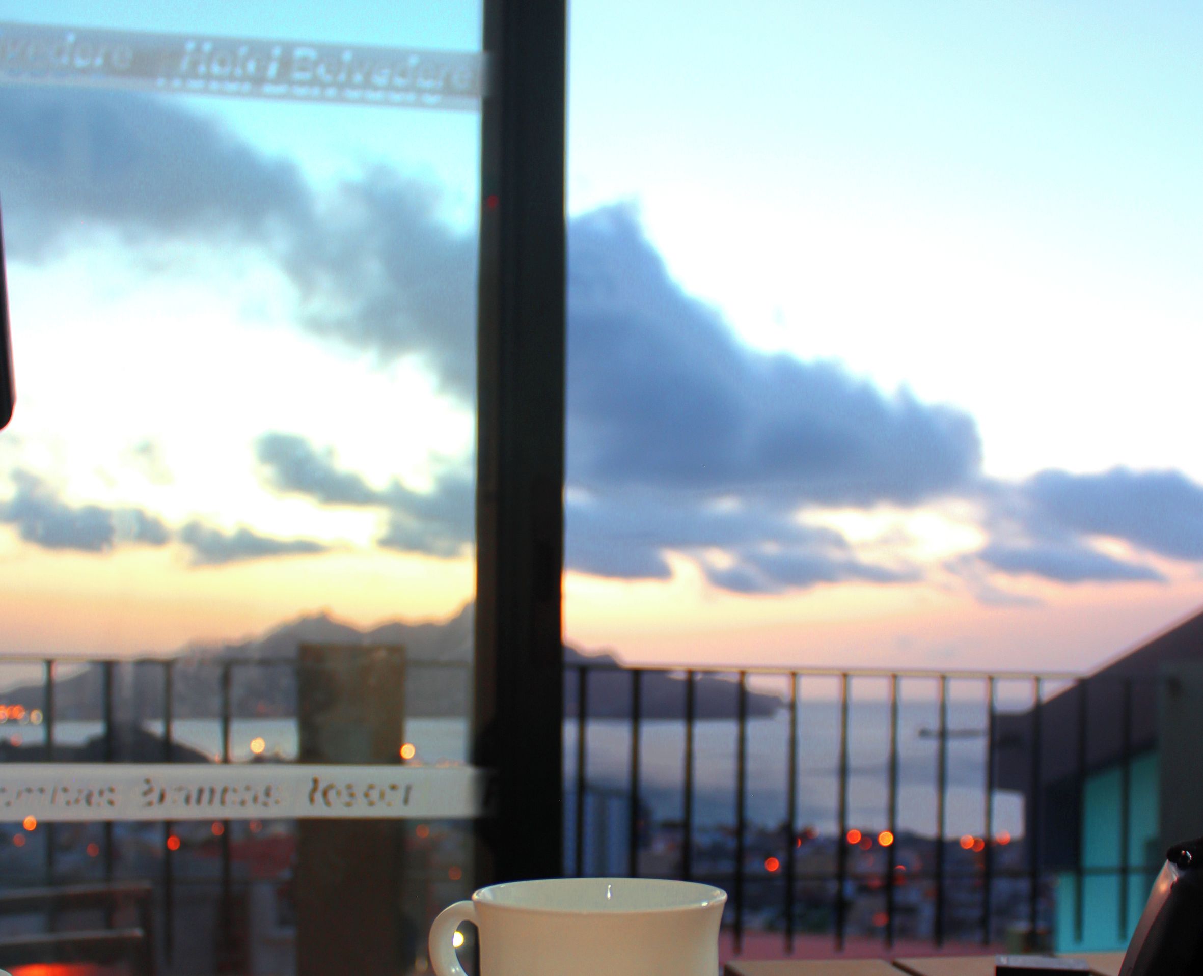 Una taza blanca con un logo sobre un platito, sobre una mesa con vista a un atardecer y nubes.