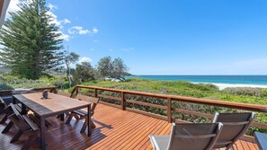 Eine Holzterrasse mit Tisch und Stühlen mit Blick auf eine Küstenlandschaft mit Bäumen und dem Meer.
