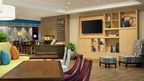 A modern hotel lobby with a sofa, dining table, chairs, a bar area, and a TV on the wall. - Home2 Suites By Hilton Cullman (Cullman)