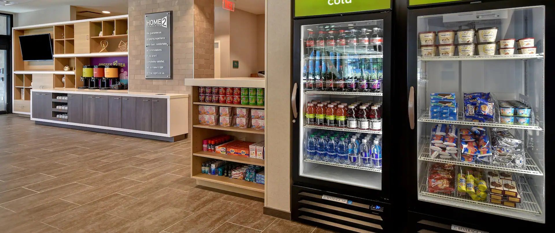 A hotel reception area with a beverage refrigerator stocked with various drinks, a coffee machine, and shelves with snacks and toiletries.