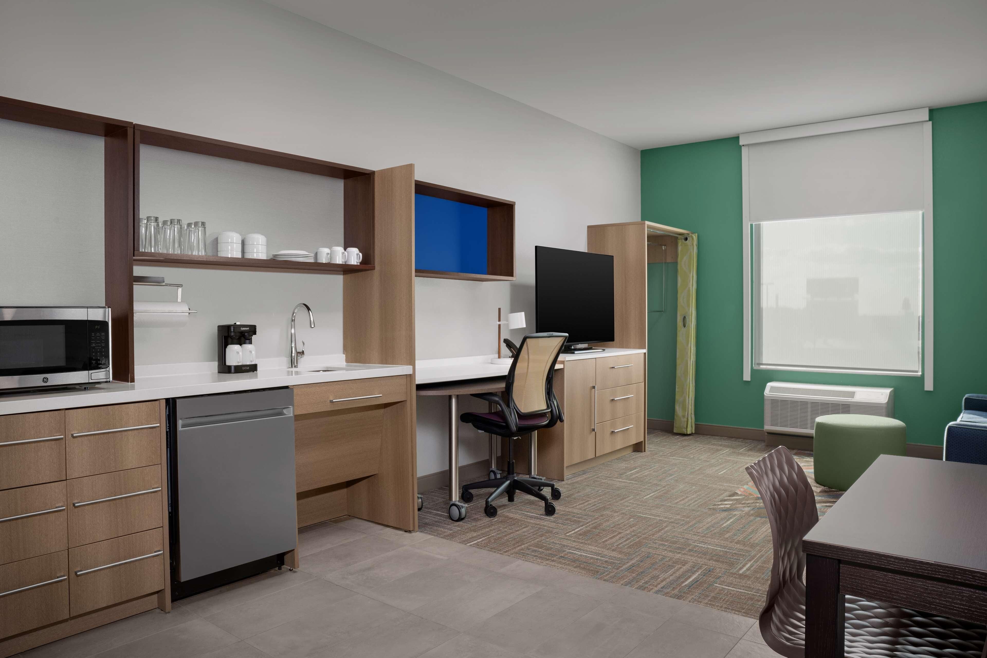 A modern kitchen with a microwave, coffee maker, and dishwasher, a dining area with a table and chairs, and a large window with blinds.