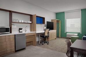A modern kitchen with a microwave, coffee maker, and dishwasher, a dining area with a table and chairs, and a large window with blinds. - Home2 Suites By Hilton Cullman (Cullman)