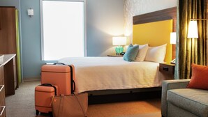 A hotel room with a bed, a desk, a chair, a suitcase, and a lamp. - Home2 Suites By Hilton Cullman (Cullman)